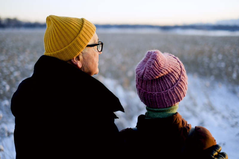 An elderly man and a woman stand with their backs to the camera, looking at a winter landscape, wearing colorful beanies.